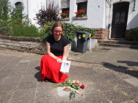 Eine Frau kniet auf dem Pflaster und hält ein Bild über einen Stolperstein mit Rosen