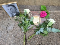 Stolperstein mit einem schwarz-weißen Porträtfoto und Rosen auf dem Boden