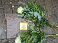 Zwei weiße Rosen liegen links und rechts neben einem Stolperstein auf grauem Pflaster